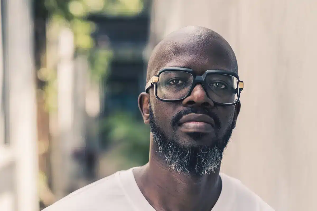 South African DJ Black Coffee stands, wearing a white tee shirt and clear glasses. 